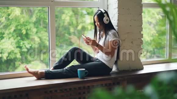 可爱的亚洲女孩深褐色头发正坐在窗台上用耳机听音乐用智能手机休息视频的预览图