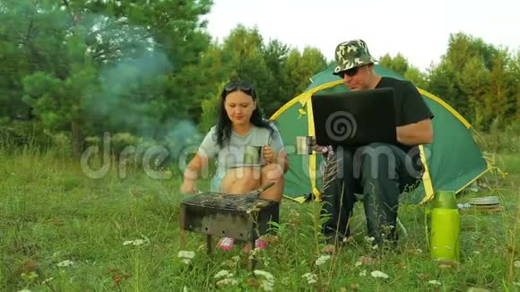 一男一女在帐篷附近的大自然中煎什串喝茶一个男人在用笔记本电脑视频的预览图