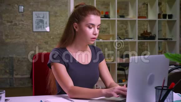一位留着红发的高加索美女一边看屏幕一边坐在室内一边用电脑视频的预览图