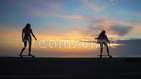 日落时分两个女孩坐在滑板上靠着岩石和天空在路上行走视频的预览图