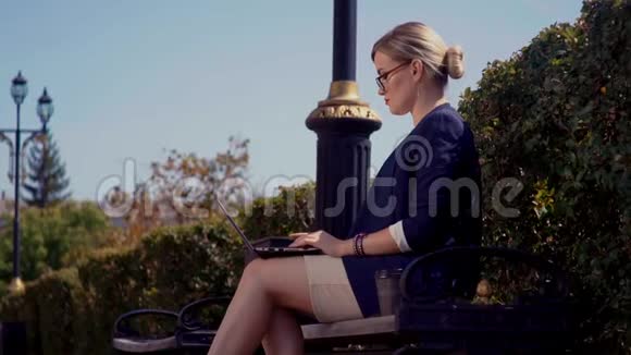 女孩坐在城市的街道上坐在长凳上她在膝上的笔记本电脑上工作在她的身后一个整齐的植物栅栏视频的预览图