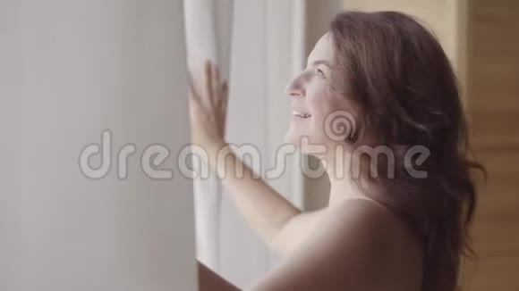 漂亮成熟的女人打开窗帘花边站在豪华公寓家或现代酒店透过窗户看视频的预览图