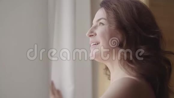 可爱的成熟女人拉开窗帘花边站在豪华公寓的家或现代酒店透过窗户欣赏视频的预览图
