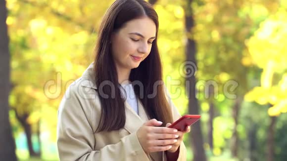 穿着外套或壕沟的年轻女子用智能手机站在秋季公园里视频的预览图