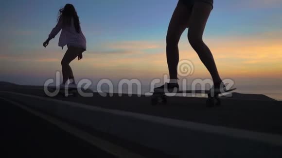 日落时分两个女孩坐在滑板上靠着岩石和天空在路上行走视频的预览图