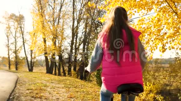 穿红色夹克的女孩正在夕阳的亮光下骑自行车在街上在黄树的背景下视频的预览图
