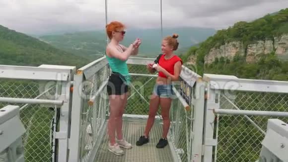 两个白种人姐妹在高山上的吊桥上笑着说话红色和金色的头发视频的预览图