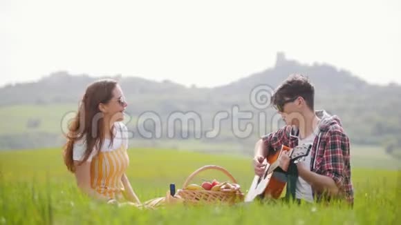 一对年轻夫妇穿着鲜艳的衣服坐在明亮的绿色草地上野餐一个男人弹吉他视频的预览图