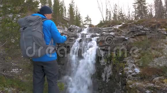 游客背着背包站在美丽的山瀑布附近他在智能手机上拍照积极主动视频的预览图