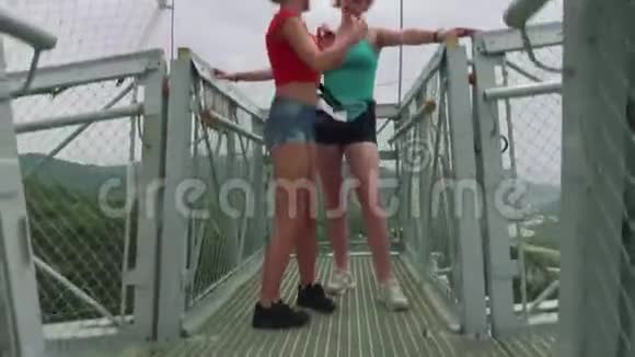 两位年轻女性自拍站在青山谷和河流背景下的悬索桥上照相机视频的预览图