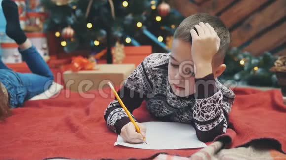 可爱的小男孩穿着节日礼服躺在地板上写信给圣诞老人圣诞树上有灯和礼物视频的预览图