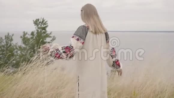 后景为敬畏的白种人女人穿着夏日的长时装站在湖边或湖边的田野上视频的预览图