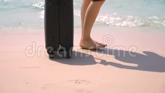特写镜头女性光着脚在海边的白色海滩上的一个手提箱旁边旅行概念视频的预览图