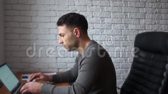 办公室里一个疲惫的年轻程序员正在用笔记本电脑工作一个人坐在皮椅上靠在一块白砖上视频的预览图