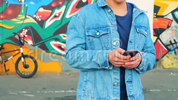 穿着牛仔裤夹克的青少年正拿着手机站在涂鸦墙附近视频的预览图