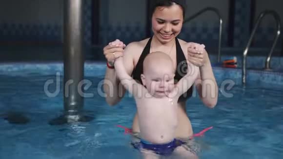 健康中心游泳池里的一位年轻母亲正和小儿子一起做水上运动视频的预览图