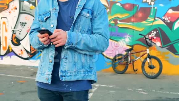 男青少年站在他的BMX和街墙附近手里拿着智能手机视频的预览图