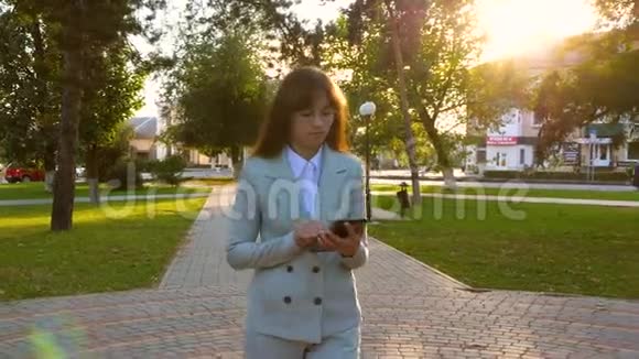 穿浅色西装的女孩手里拿着平板电脑上班美丽的女商人在公园的小巷里散步并检查视频的预览图
