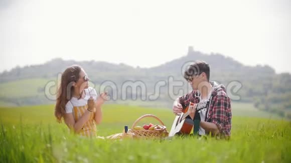 一对年轻夫妇穿着鲜艳的衣服坐在明亮的绿色草地上野餐一个男人弹吉他一个女人视频的预览图