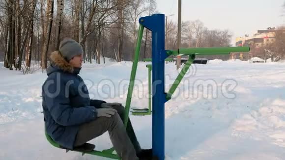 穿着带罩的蓝羽绒服的人正在冬季公园的模拟器上做腿部运动侧视视频的预览图