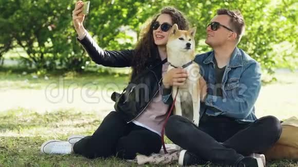 快乐的一对情侣正和他们的狗在公园的草地上玩智能手机自拍人们摆姿势说话视频的预览图