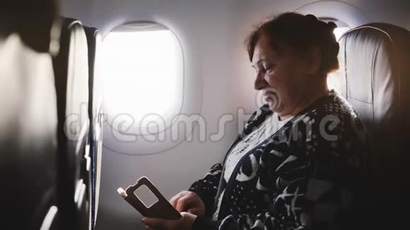 快乐高级女性飞机乘客使用智能手机信使应用程序享受风景和微笑的窗口座位视频的预览图