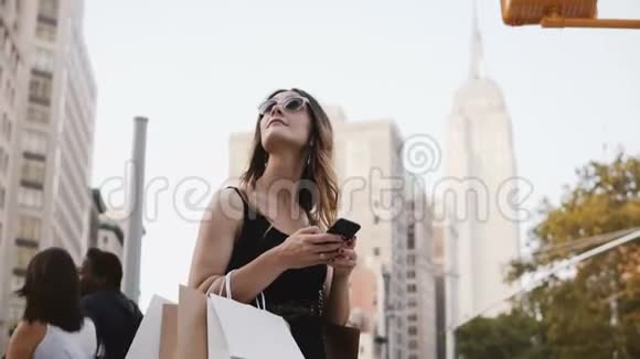 快乐迷人的白种人年轻女性带着太阳镜的购物袋向上看在纽约使用智能手机应用程序视频的预览图