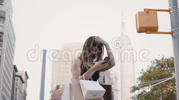 快乐的欧洲女性自由职业者带着太阳镜购物袋微笑使用智能手机应用程序在纽约市视频的预览图