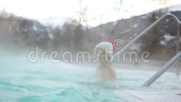 穿着一顶带红色波姆的针织灰色帽子和一件泳衣的美丽的黑发女人在温泉里游泳视频的预览图