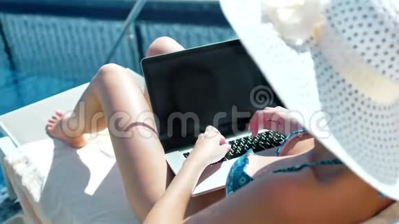 穿着泳衣戴着太阳帽的女人在游泳池附近的躺椅上用笔记本电脑写文字视频的预览图