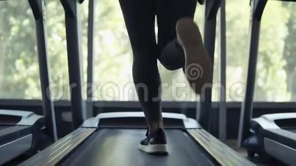 穿着黑色胸罩和灰色紧身裤的女人在健身房全景窗口前的跑步机上跑步背面视图视频的预览图