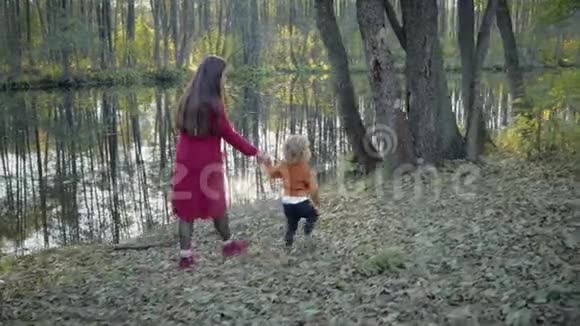 母亲和儿子穿着时髦的衣服走在秋天的森林里在湖边树木的倒影可以看到视频的预览图