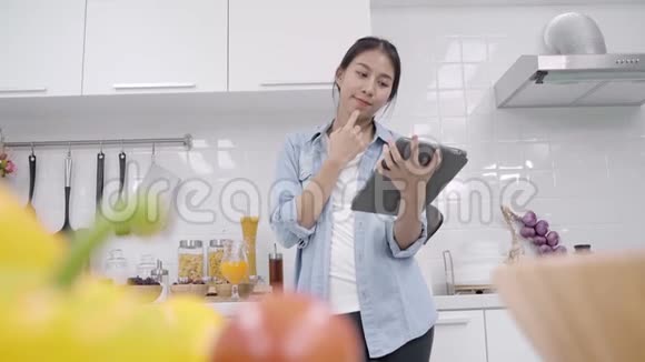 快乐的亚洲女人在厨房里做食物时用平板电脑看食谱而女性则用有机蔬菜来健康视频的预览图
