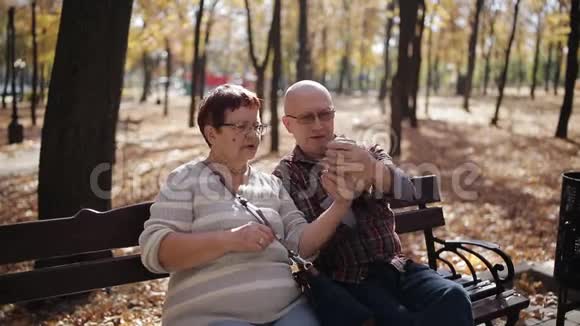 秋天公园的一对老夫妇坐在长凳上用智能手机自拍视频的预览图