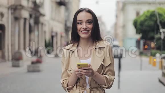 微笑的美丽少女手持手机站在镜头前微笑着看城市背景的画像视频的预览图