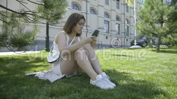 阳光明媚的一天一个年轻漂亮的女人盘腿坐在公园的草地上用智能手机工作视频的预览图