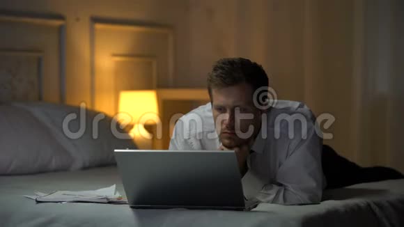 精疲力竭的工程师躺在床上工作在笔记本电脑和偏航阅读邮件视频的预览图