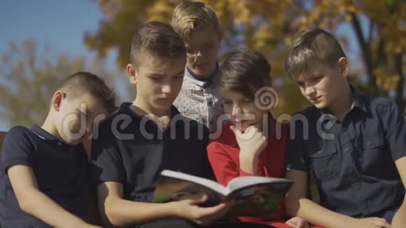 男孩们一起坐在长凳上骑着一本书朋友们在秋天的公园里度过一个阳光明媚的日子视频的预览图