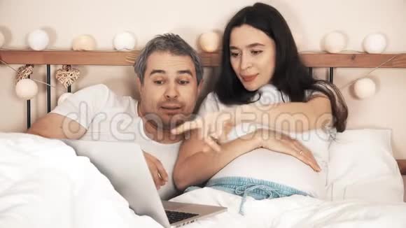 快乐的男人和怀孕的妻子在家里的床上用笔记本电脑视频的预览图
