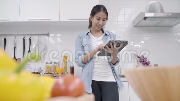快乐的亚洲女人在厨房里做食物时用平板电脑看食谱而女人则用有机蔬菜做健康的食物视频的预览图