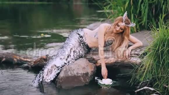 一头金发碧眼的海妖头上戴着一顶美人鱼珍珠王冠头上戴着一条豪华的银色尾巴坐在原木上视频的预览图