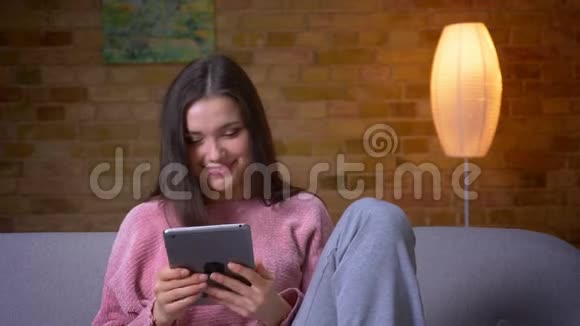 一位年轻漂亮的棕色白种人女性使用平板电脑在一个舒适的沙发上微笑着特写镜头视频的预览图