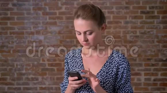 可爱的白种人女性轻轻地挥动她的手机自信地看着镜头在砖背景上视频的预览图
