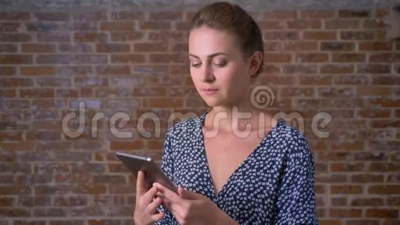 漂亮的白种人年轻女子在平板电脑上显示绿色屏幕站在砖墙旁边高兴地眨着眼睛视频的预览图