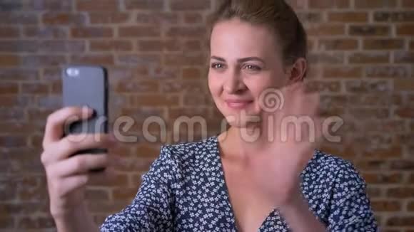 美丽的白种人黑发女孩正在进行视频通话并在红砖旁边微笑和欢乐地挥舞着她的智能手机视频的预览图