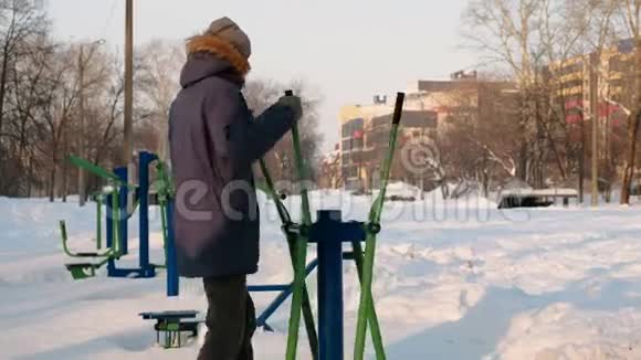 穿着带罩的蓝羽绒服的人在一个冬季城市公园里从事滑雪模拟器后景视频的预览图