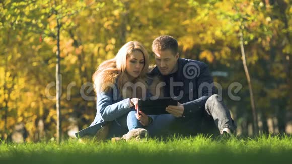情侣在网上购物在秋季公园使用平板电脑在网上购买保暖衣服视频的预览图