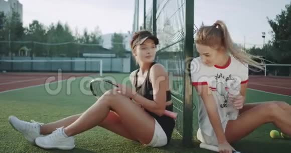 两位漂亮的女士在网球赛后坐在地板上休息喝一瓶水视频的预览图