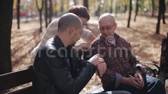 年长的父母和成年的儿子在秋天公园的长凳上交流老人向父母解释如何使用视频的预览图