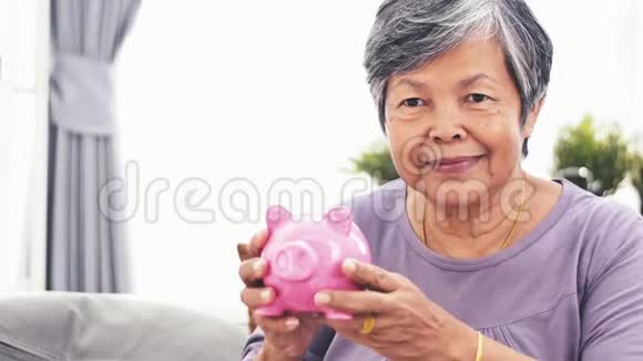 一位兴高采烈的亚洲老太太对着镜头微笑着摇着存钱罐成年女子举着储蓄罐存钱的画像印视频的预览图